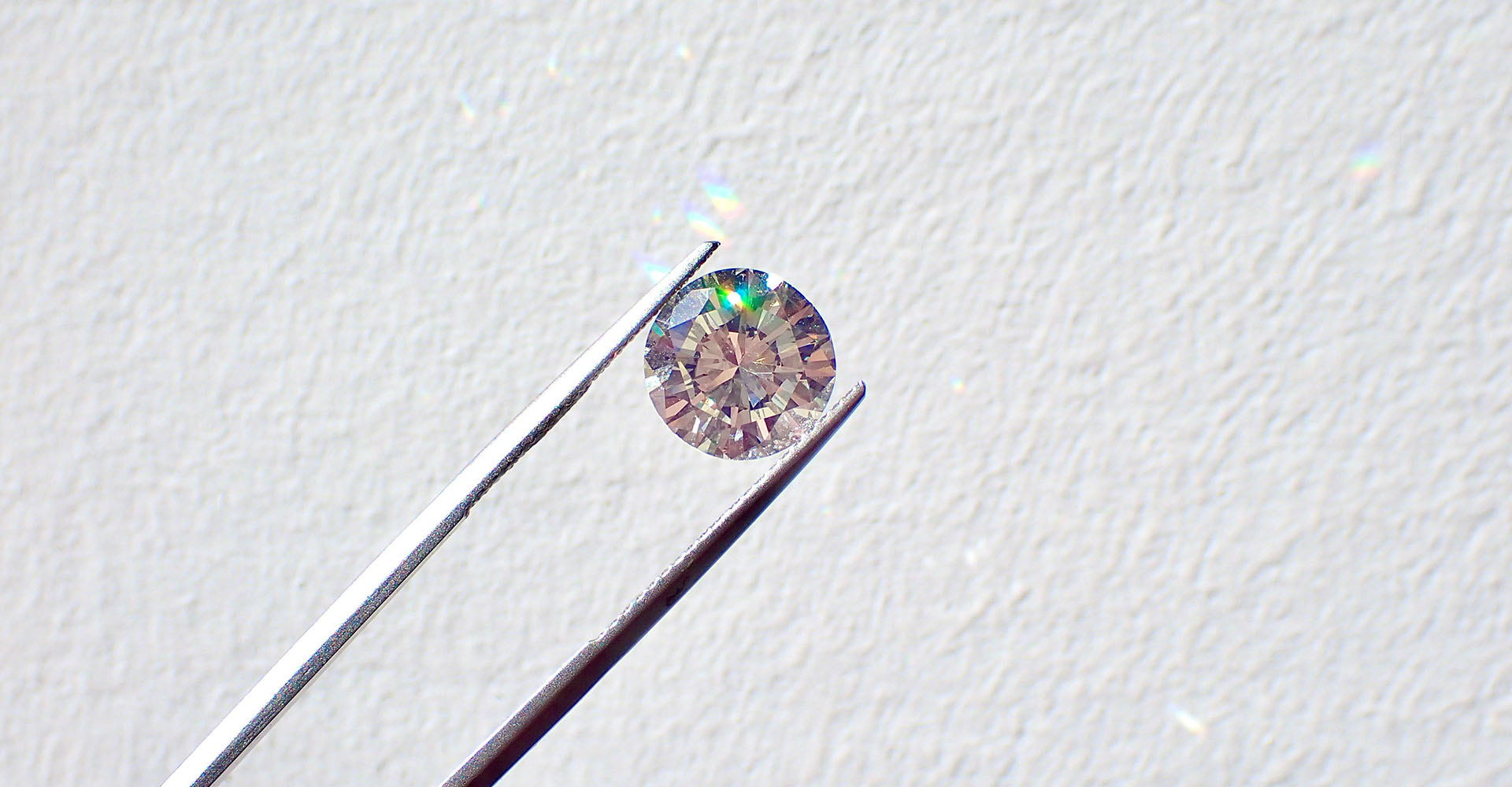 close up of tongs holding up diamond against a white wall, rainbow light reflecting on the white wall, Sheaff Brock investment advisors blog, retail consumers